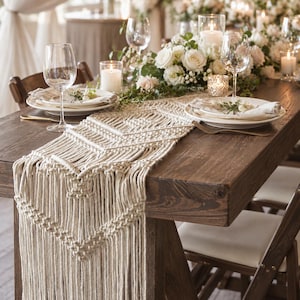May include: A cream-colored macrame table runner featuring intricate knotted geometric patterns and long fringe edges, draped over a dark brown wooden table set with white ceramic plates, clear wine glasses, and lit pillar candles.