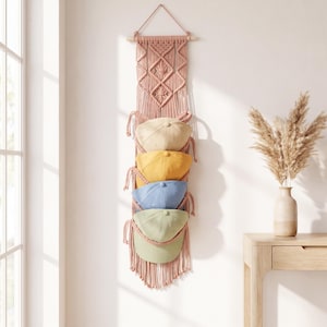 May include: A vertical macrame wall hanging in a dusty rose colour featuring intricate knotted patterns and four integrated loops designed to hold baseball caps. The organiser displays four baseball caps in beige, mustard yellow, light blue, and sage green.
