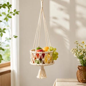 Puede incluir: Una cesta de frutas colgante de macramé llena de manzanas, uvas y limones. La cesta está hecha de algodón tejido de color crema y tiene una borla decorativa en la parte inferior. La cesta está suspendida de un solo lazo en la parte superior.