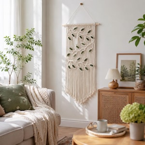 May include: A cream-colored macrame wall hanging suspended from a wooden dowel, featuring a woven branch design with dark green leaf accents. The piece consists of long, vertical knotted cords that form a textured, trailing vine pattern.