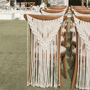 Pode incluir: Fileiras de cadeiras de madeira com decorações de macramé em um gramado. As cadeiras têm almofadas de cores claras e são montadas para um evento ao ar livre. O macramé adiciona um toque boêmio.