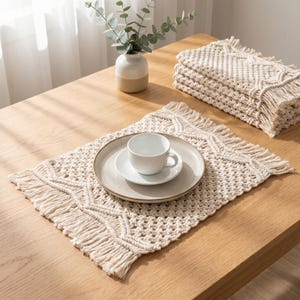 Puede incluir: Un mantel individual de macramé beige con un diseño geométrico y flecos. Una taza y un platillo blancos descansan sobre un plato beige. Una pila de manteles individuales a juego y un jarrón con vegetación están sobre una mesa de madera.