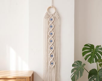 May include: Cream-colored macrame wall hanging with a wooden ring at the top. The design features a series of blue and white evil eye beads. The bottom has a fringe. The wall hanging is displayed against a white wall.