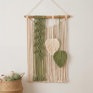 May include: Macrame wall hanging featuring a natural wood dowel and cream-colored cotton cords. The design includes a vertical panel of olive green macrame with leaf details, and two leaf-shaped accents in cream and green.