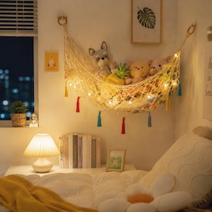 May include: A cream-colored macrame toy hammock suspended by two wooden rings, featuring a woven net design with hanging tassels in red, yellow, and teal. The hammock is illuminated by a string of small star-shaped lights and holds several plush toys.