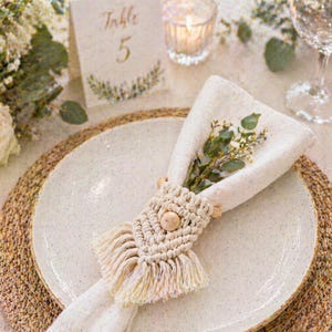 May include: A table setting with a white plate, napkin, and macrame napkin ring. The napkin is adorned with greenery. A gold fork and knife are on either side of the plate. A table number card reads "Table 5".