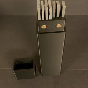 Peut inclure: Un ensemble de blocs de couteaux noirs de forme rectangulaire. L'ensemble comprend un grand bloc rectangulaire avec des fentes à couteaux grises et un petit récipient carré pour accessoires. Les fentes à couteaux sont remplies de couteaux gris.