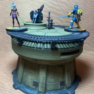 Op de afbeelding: Gedetailleerd miniatuur diorama met twee actiefiguren op een platform met meerdere niveaus. Eén figuur houdt een groen lichtzwaard vast, de ander een blaster. Het platform is voornamelijk olijfgroen met zwarte en grijze accenten, wat wijst op een sci-fi- of fantasiethema.