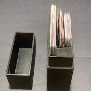 Peut inclure: Deux boîtes noires rectangulaires pour cartes. L'une est vide, l'autre contient une pile de cartes. Les cartes sont visibles et présentent une variété de couleurs. Les boîtes semblent être en plastique.