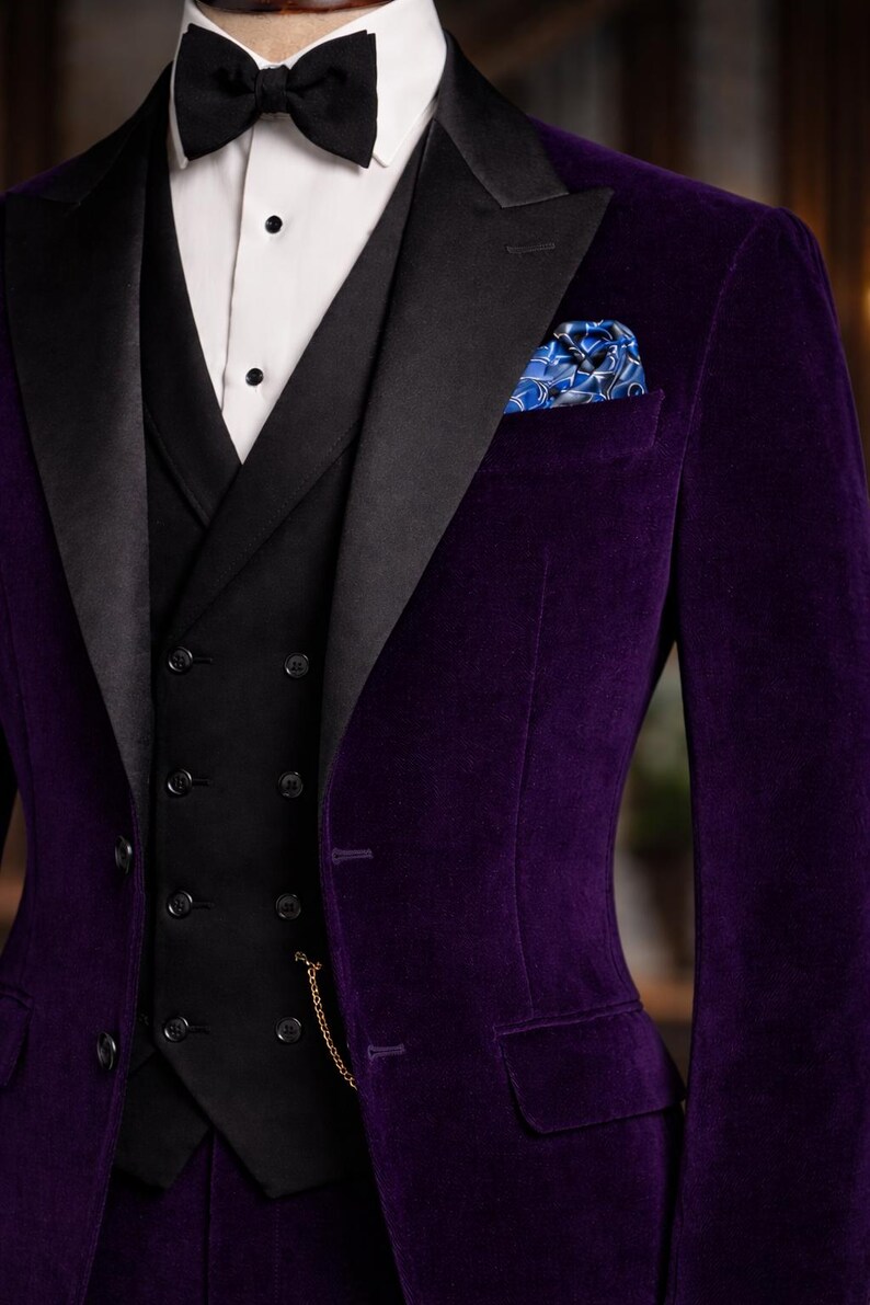Puede incluir: Un conjunto formal con una blazer de terciopelo morado oscuro con solapas negras, un chaleco negro y una camisa blanca. Una pajarita negra y un pa&ntilde;uelo de bolsillo estampado a&ntilde;aden detalles. El traje tiene un corte cl&aacute;sico y entallado.
