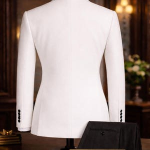 Pode incluir: Um casaco de fato branco exposto num manequim, com cal&ccedil;as pretas. O casaco tem um design cl&aacute;ssico com quatro bot&otilde;es em cada punho. Uma placa dourada na base diz "The Noble Tailor".