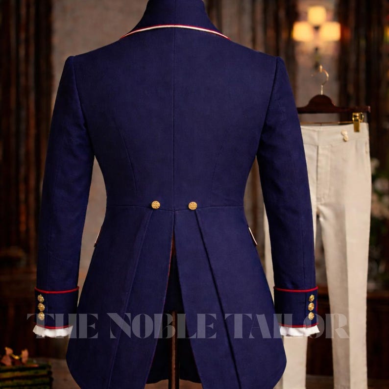K&ouml;nnte beinhalten: Marineblaue Jacke im Milit&auml;rstil mit roter und wei&szlig;er Verzierung, goldenen Kn&ouml;pfen und geteiltem R&uuml;cken. Die Jacke hat lange &Auml;rmel und einen hohen Kragen. Der Text "THE NOBLE TAILOR" ist unten sichtbar. Eine cremefarbene Hose h&auml;ngt im Hintergrund.