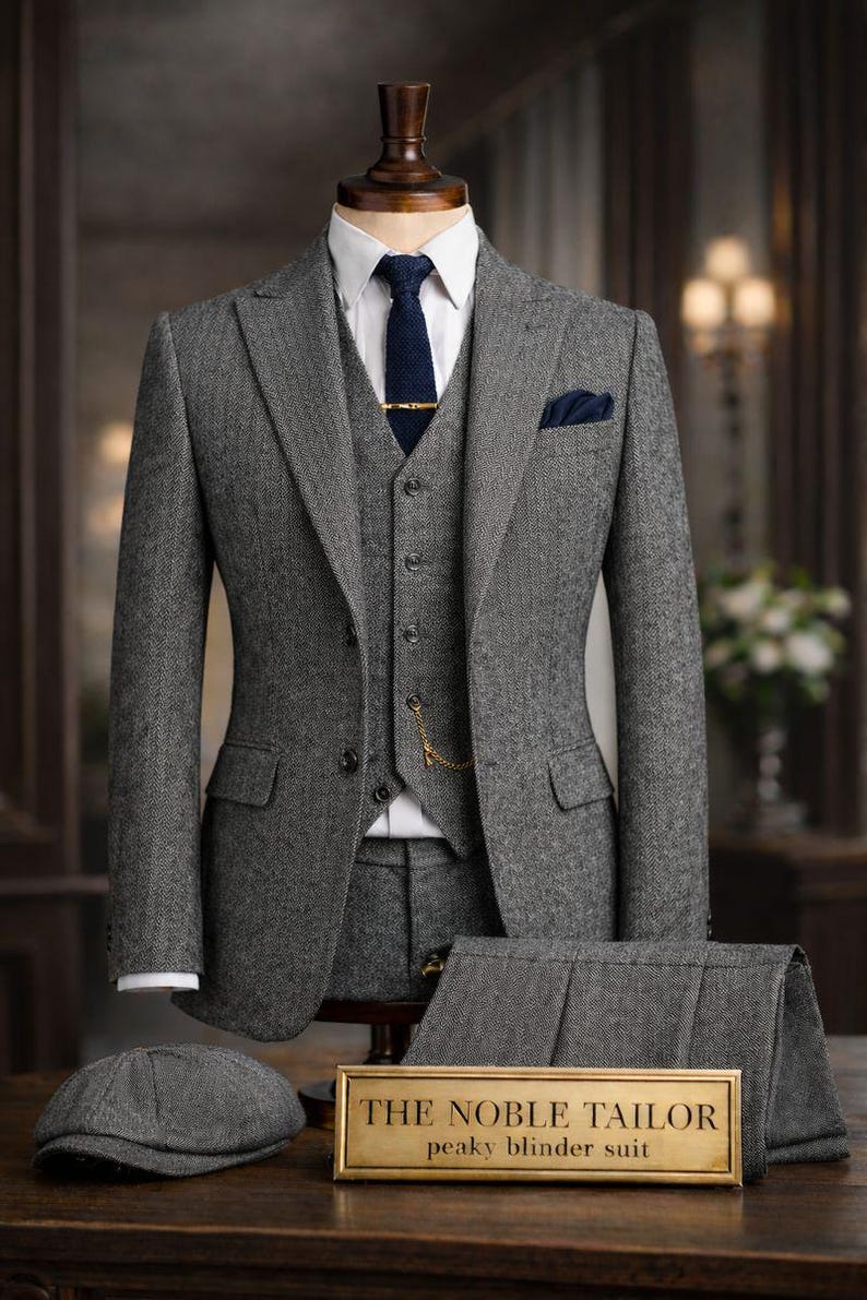 May include: A gray herringbone three-piece suit with a matching flat cap and trousers, displayed on a mannequin. The suit includes a white dress shirt, navy tie, and pocket square. A gold sign reads "THE NOBLE TAILOR" and "peaky blinder suit."