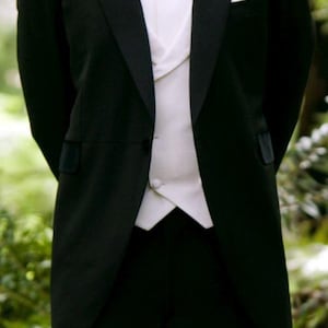 Può includere: Uno smoking nero formale con gilet, camicia e papillon bianchi. Un fiore bianco è appuntato sulla bavero. La giacca ha un colletto di velluto e un fazzoletto da taschino. L'outfit è adatto per un matrimonio o un evento formale.