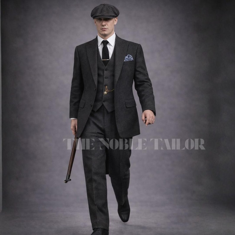 May include: A charcoal gray three-piece suit with a matching flat cap. The suit includes a blazer, vest, and trousers, paired with a white dress shirt and black tie. A pocket square and a pocket watch add detail. The text "THE NOBLE TAILOR" is visible.