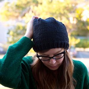 Op de afbeelding: Een persoon draagt een donkergroene trui en een zwarte gebreide beanie met een rand. De persoon draagt een bril en heeft lang bruin haar.