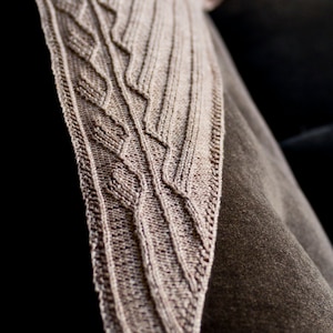 Könnte beinhalten: Nahaufnahme einer gestrickten Stola in beige mit einem strukturierten Muster. Die Stola liegt auf einer braunen Oberfläche.