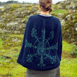 Puede incluir: Un poncho de punto azul oscuro con un diseño de copo de nieve turquesa en la espalda. El poncho lo lleva puesto una persona de pie en un campo de hierba.