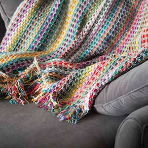 May include: A multi-coloured, hand-woven blanket with a fringed edge. The blanket displays a vibrant mix of colours including red, blue, green, yellow, and pink. The blanket is draped over a grey sofa, showcasing its texture and design.
