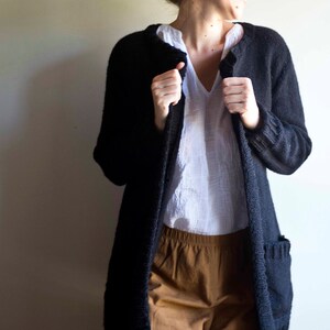 Op de afbeelding: Een vrouw draagt een zwarte cardigan over een wit shirt en een bruine broek. Ze glimlacht en kijkt naar de zijkant.