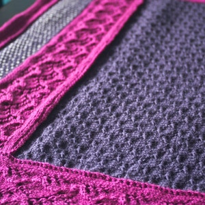 May include: A knitted blanket with a purple center and a pink border. The blanket is made with a textured stitch pattern.