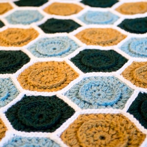 May include: A crocheted blanket with a honeycomb pattern in shades of green, blue, and brown. The blanket is made with a variety of stitches and textures, creating a unique and interesting design.