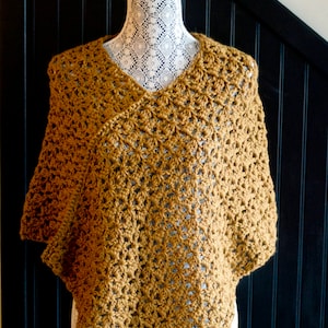 Może przedstawiać: Ponczo wykonane na szydełku w kolorze musztardowym. Ponczo jest prezentowane na białym manekinie z koronkowymi ozdobami.