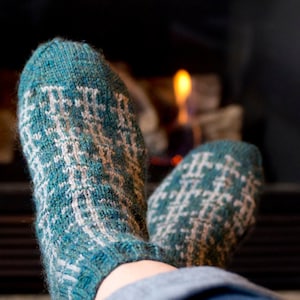 Op de afbeelding: Een paar gebreide sokken met een turquoise en wit patroon. De sokken hebben een geometrisch ontwerp en worden gedragen bij een open haard. De sokken zijn gemaakt van een zacht, warm materiaal, ideaal voor koud weer.