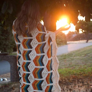May include: A person wearing a colourful crocheted blanket with a geometric pattern, standing in front of a sunset.
