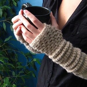 Könnte beinhalten: Ein Paar beigefarbene Fingerlose-Handschuhe aus Häkelarbeit mit einem strukturierten Muster. Die Handschuhe werden von einer Person getragen, die eine dunkelbraune Tasse hält.