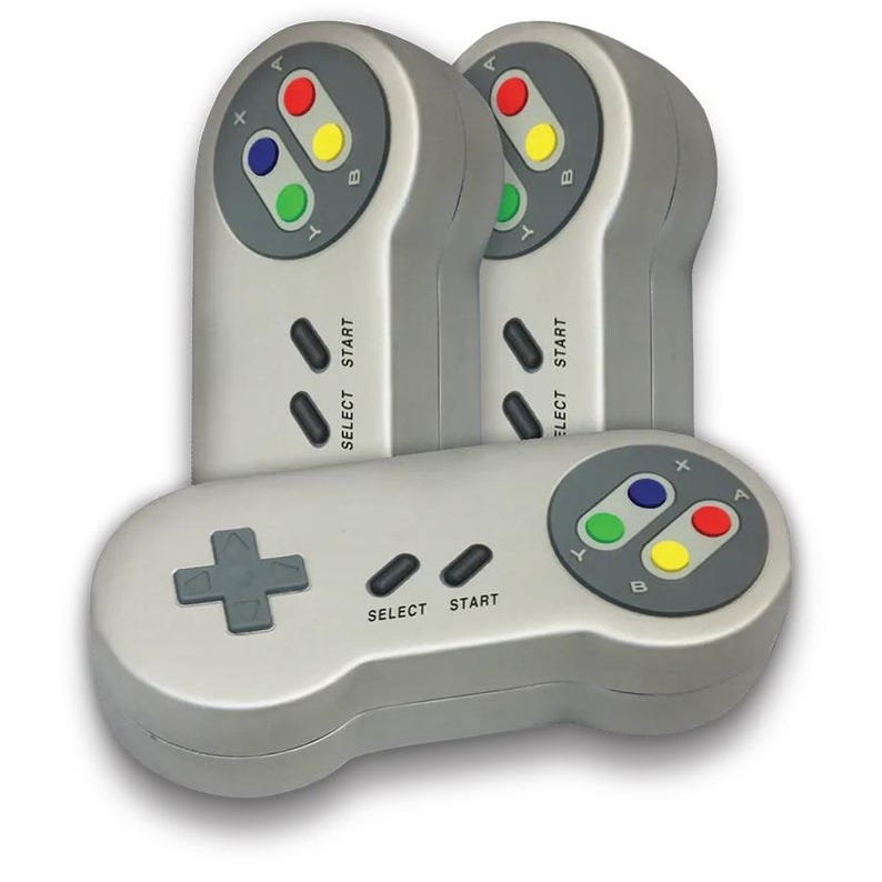 May include: Three gray retro-style video game controllers. Each controller has a directional pad, select and start buttons, and colored buttons. The controllers are designed to resemble a classic console controller.