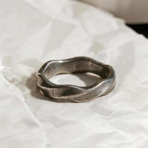 May include: A silver-toned ring with a wavy, textured design. The ring is set against a white, crumpled paper background. The metal has a slightly tarnished appearance, suggesting an aged or vintage aesthetic. The ring's shape is irregular, adding to its unique appeal.