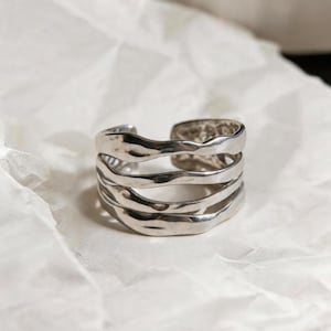 Puede incluir: Un anillo de puño plateado con un diseño texturizado y en capas. El anillo presenta cuatro bandas de metal ondulado e irregular, creando una estética única y moderna. El anillo está sobre un fondo blanco texturizado.