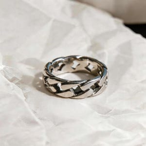 Puede incluir: Un anillo plateado con un diseño de eslabones de cadena. El anillo está hecho de un metal brillante y descansa sobre una superficie blanca texturizada. El diseño del anillo presenta eslabones interconectados, creando un aspecto audaz y moderno.