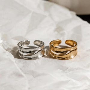 Puede incluir: Dos anillos abiertos, uno plateado y otro dorado, descansan sobre una superficie blanca texturizada. Cada anillo presenta un diseño en capas con un patrón ondulado y texturizado. Los anillos son probablemente ajustables.