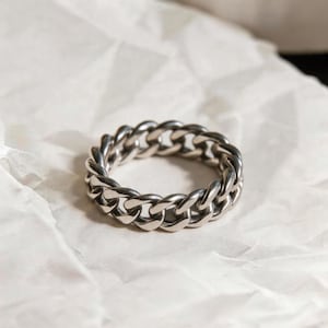 Puede incluir: Un anillo plateado con un diseño de eslabones de cadena. El anillo, hecho de metal brillante, descansa sobre una superficie blanca texturizada. Los eslabones están entrelazados, formando una banda continua.