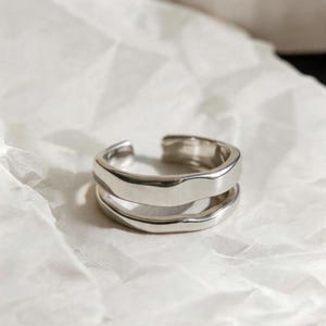 Puede incluir: Un anillo plateado con un diseño único en capas. El anillo presenta dos bandas con una superficie ondulada y texturizada. El diseño abierto permite un ajuste ajustable. La joya se exhibe sobre una superficie blanca y texturizada.