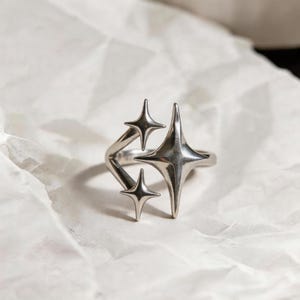 Puede incluir: Un anillo plateado con un diseño de cuatro estrellas. El anillo se encuentra sobre un fondo blanco texturizado. Las estrellas tienen una superficie pulida y reflectante, creando una estética moderna y celestial.