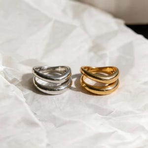 Puede incluir: Dos anillos, uno plateado y otro dorado, se exhiben sobre una superficie blanca texturizada. Cada anillo presenta un diseño de doble banda, con una apariencia suave y redondeada. Los anillos están colocados uno al lado del otro, mostrando sus colores contrastantes y estilos similares.