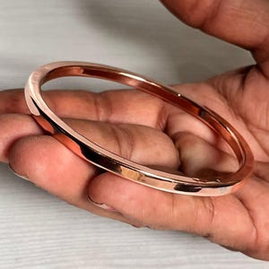 Op de afbeelding: Een gepolijste koperkleurige armband. De gladde, afgeronde armband wordt in een hand gehouden en toont zijn reflecterende oppervlak. De armband is een eenvoudig, elegant ontwerp, geschikt voor dagelijks gebruik.