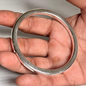 Può includere: Un braccialetto a cerchio argentato lucido. Il braccialetto rotondo ha una superficie liscia ed è tenuto in una mano. Il design è semplice ed elegante. Questo pezzo è adatto all'uso quotidiano. Il diametro del braccialetto è di circa 6,5 cm.