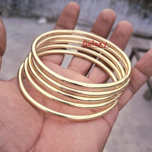 May include: A stack of gold-colored bangles, with a smooth, polished finish. The bangles are round and appear to be made of metal. The word "Galaxy" is written in red on the bangles. The bangles are held in a person's hand.