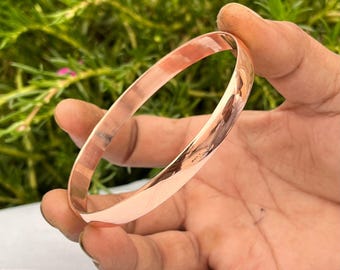 May include: A polished copper bangle bracelet. The bracelet is a solid band with a smooth, reflective surface. The copper has a warm, reddish-brown hue. The bracelet is held in a hand against a blurred green background.