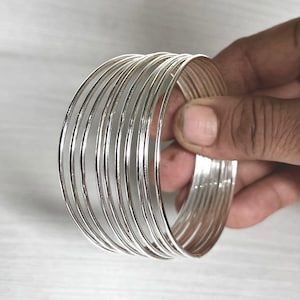 Puede incluir: Un brazalete plateado con múltiples bandas finas. El diseño abierto se muestra con una mano, resaltando el brillo del metal pulido. Este brazalete es un accesorio de moda, adecuado para diversas ocasiones.
