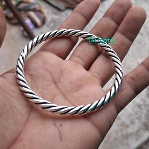 Peut inclure: Un bracelet jonc circulaire en argent présentant un motif de corde torsadée en continu. La surface métallique a une finition polie avec des accents oxydés sombres dans les rainures pour souligner la texture en spirale. Le mot Drishti est écrit en sarcelle.