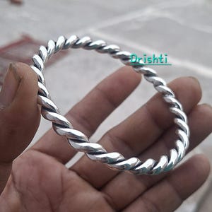 Puede incluir: Un brazalete de color plateado pulido con un diseño de cuerda trenzada continua. La superficie metálica tiene un acabado reflectante con una sutil oxidación oscura en las ranuras. La palabra Drishti está impresa en color turquesa arriba.