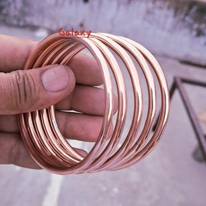 May include: A stack of rose gold bangles. The smooth, circular bracelets are held in a hand. The word "Galaxy" is written in red script across the top of the image.