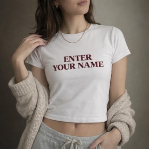 May include: White crop top with the text "ENTER YOUR NAME" in burgundy. The shirt has short sleeves and is paired with gray sweatpants and a beige cardigan. A gold necklace is also visible.