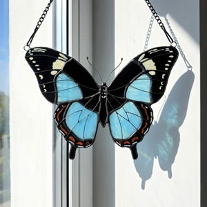 May include: A stained glass butterfly with black, light blue, and white wings, hanging from a black chain. The butterfly's wings have a detailed design with black outlines and orange accents. The butterfly is casting a shadow on a white surface.