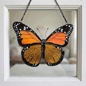 K&ouml;nnte beinhalten: Glasmalerei Monarchfalter mit orangefarbenen und braunen Fl&uuml;geln, schwarzen Umrissen und wei&szlig;en Punkten. Der Schmetterling wird von einer schwarzen Kette gehalten und in einem wei&szlig; gerahmten Fenster ausgestellt. Dekoratives Kunstwerk.
